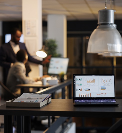 Laptop computer with financial graphs on screen, standing on table in startup office, coworkers discussing company strategy in background. Employees working late at night in startup office
